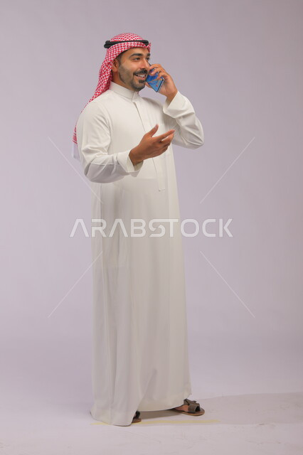 تبادل الاحاديث المسلية والحوارات، التسامر مع الأصدقاء والأقارب بإيماءات السعادة، مفهوم صلة الرحم، بورتريه لرجل عربي خليجي سعودي يرتدي الثوب والشماغ التقليدي يقوم بإجراء مكالمة صوتيه عبر الهاتف المحمول، صورة الجسم بالطول الكامل، خلفية رمادية
