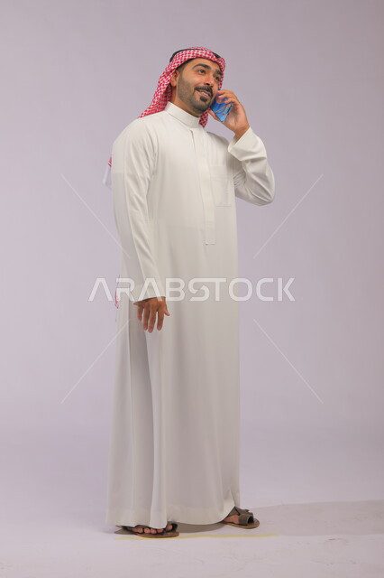 مفهوم صلة الرحم، تبادل الاحاديث والحوارات، التسامر مع الأصدقاء والأقارب بإيماءات السعادة، بورتريه لرجل عربي خليجي سعودي يرتدي الثوب والشماغ التقليدي يقوم بإجراء مكالمة صوتيه عبر الهاتف المحمول، صورة الجسم بالطول الكامل، خلفية رمادية