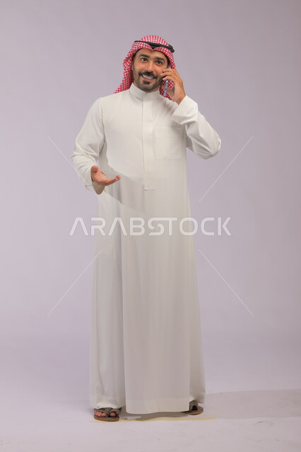 تبادل الاحاديث والحوارات، التسامر مع الأصدقاء والأقارب بإيماءات السعادة، مفهوم صلة الرحم، بورتريه لرجل عربي خليجي سعودي يرتدي الثوب والشماغ التقليدي يقوم بإجراء مكالمة صوتيه عبر الهاتف المحمول، صورة الجسم بالطول الكامل، خلفية رمادية
