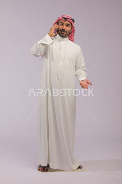 حركات تدل على الاستفسار عن شيء ما، البحث عن عمل عبر وسائل التواصل الاجتماعي, دمج التقنية والتكنولوجيا في الحياة العملية، بورتريه لرجل عربي خليجي سعودي مبتسم يرتدي الشماغ و الثوب التقليدي ويقوم بإجراء مكالمة هاتفية عبر الجوال، صورة الجسم بالكامل, خلفية رمادية