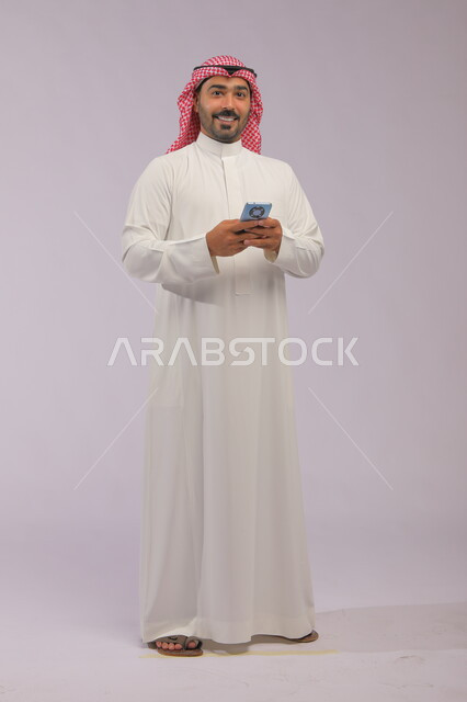 Exchanging conversations with friends via mobile applications, a portrait of a Saudi Gulf Arab young man wearing a shemagh and traditional dress, holding a mobile phone in his hand and looking at the camera, browsing the Internet and completing tasks remotely, integrating technology and technology into practical life, full-body portrait, gray background