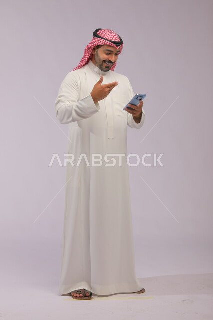إجراء مكالمة فيديو مع الأهل عبر الانترنت، تصفح تطبيقات الهاتف المحمول، بورتريه لشاب عربي خليجي سعودي مبتسم يرتدي الشماغ والثوب التقليدي يقوم بمتابعة اخبار وسائل التواصل الاجتماعي باستخدام الجوال، ايماءات الاندماج والتركيز، صورة الجسم بالكامل، خلفية رمادية