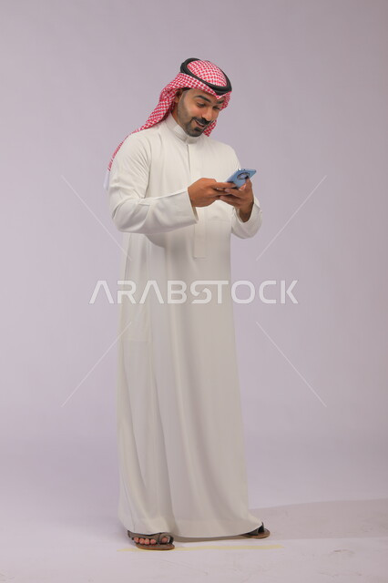 تصفح تطبيقات الهاتف المحمول، الدردشة والتحدث مع الاصدقاء عبر الانترنت، بورتريه لشاب عربي خليجي سعودي يرتدي الشماغ والثوب التقليدي يقوم بمتابعة اخبار وسائل التواصل الاجتماعي باستخدام الجوال، ايماءات الاندماج والتركيز، صورة الجسم بالطول الكامل، خلفية رمادية