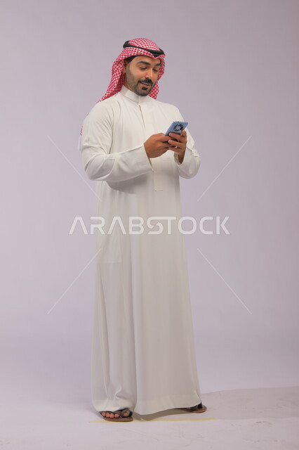  الدردشة والتحدث مع الاصدقاء عبر الانترنت، تصفح تطبيقات الهاتف المحمول، بورتريه لشاب عربي خليجي سعودي يرتدي الشماغ والثوب التقليدي يقوم بمتابعة اخبار وسائل التواصل الاجتماعي باستخدام الجوال، ايماءات الاندماج والتركيز، صورة الجسم بالطول الكامل، خلفية رمادية