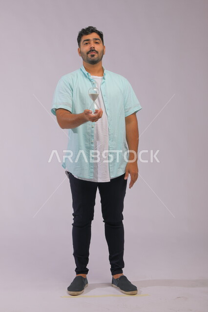 Looking at the camera with waiting gestures, setting work dates and times, monitoring sand time, the concept of business management and organizing the schedule, portrait of a Saudi Gulf man wearing casual clothes holding a glass hourglass (bankam) in his hand, full-length body portrait, gray background