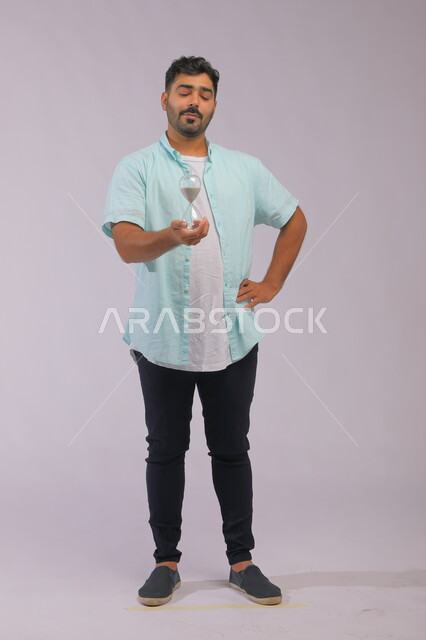 تحديد مواعيد وأوقات العمل، القيام بمراقبة توقيت الرمل, مفهوم إدارة الأعمال وتنظيم الجدول الزمني، بورتريه لرجل سعودي خليجي يرتدي الزي الكاجوال يحمل بيده الساعة الرملية الزجاجية (البنكام) وينظر إليها بإيماءات الانتظار، صورة الجسم بالطول الكامل، خلفية رمادية