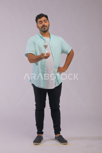 القيام بمراقبة توقيت الرمل, مفهوم إدارة الأعمال وتنظيم الجدول الزمني، بورتريه لرجل سعودي خليجي يرتدي الزي الكاجوال يحمل بيده الساعة الرملية الزجاجية (البنكام) وينظر إليها بإيماءات الانتظار، تحديد مواعيد وأوقات العمل، صورة الجسم بالطول الكامل، خلفية رمادية