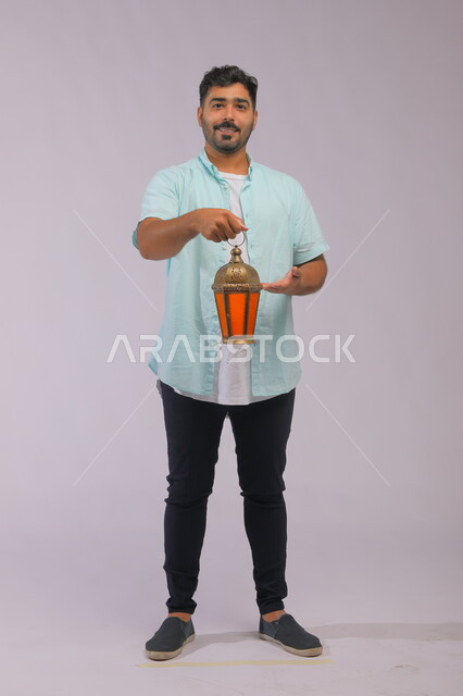 اسلاميات وعبادة, تحف وزينة الاحتفالات في السعودية, الاستعداد لقدوم شهر رمضان, المناسبات الدينية والأعياد الإسلامية, بورتريه لشاب عربي خليجي سعودي مبتسم يرتدي الزي الكاجوال يشير بيده على الفانوس المضيء, صورة الجسم بالطول الكامل, خلفية رمادية