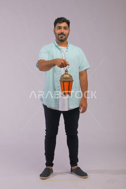 الاستعداد لقدوم شهر رمضان, المناسبات الدينية والأعياد الإسلامية, بورتريه لشاب عربي خليجي سعودي مبتسم يرتدي الزي الكاجوال يحمل بيده الفانوس المضيء, تحف وزينة الاحتفالات في السعودية, صورة الجسم بالكامل, اسلاميات وعبادة, خلفية رمادية