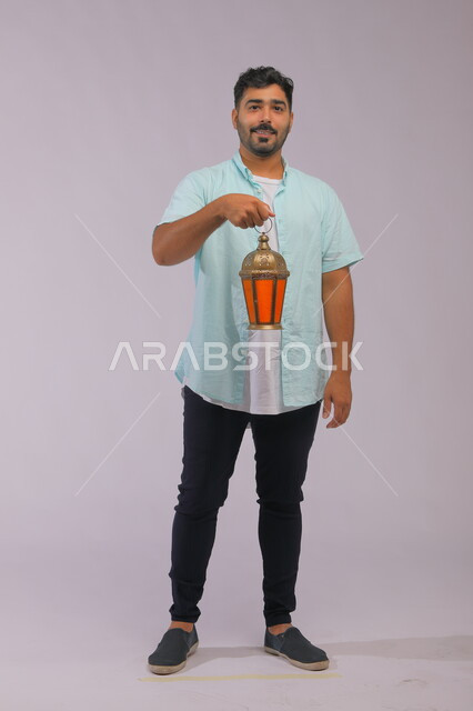  تحف وزينة الاحتفالات في السعودية, الاستعداد لقدوم شهر رمضان, المناسبات الدينية والأعياد الإسلامية, بورتريه لشاب عربي خليجي سعودي مبتسم يرتدي الزي الكاجوال يحمل بيده الفانوس المضيء, صورة الجسم بالكامل, اسلاميات وعبادة, خلفية رمادية