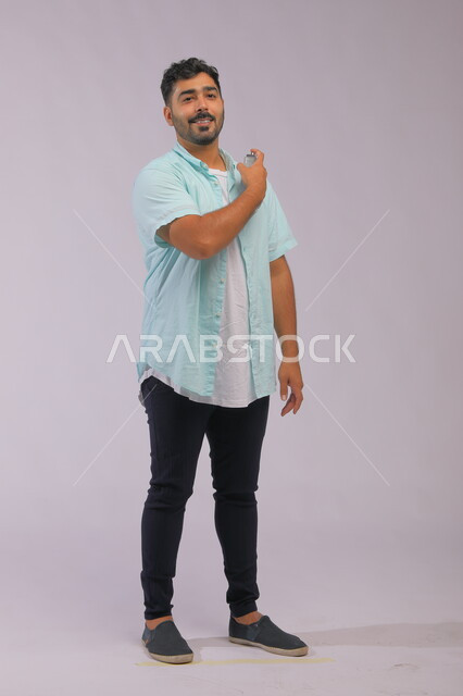  التسويق لمنتجات العطور, مفهوم التعطر والتطيب، عطورات خليجية سعودية, الاهتمام بالمظهر الخارجي, استخدام الزيوت العطرية الطبيعية, بورتريه لشاب عربي خليجي سعودي يرتدي الزي الكاجوال و يقوم برش العطر , صورة الجسم بالكامل, خلفية رمادية