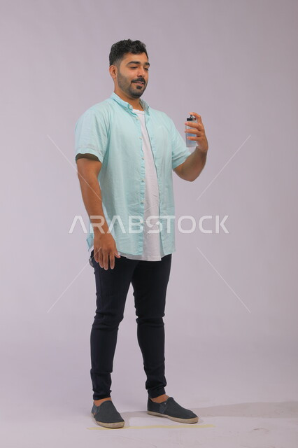 مفهوم التطيب والتعطر، عطورات خليجية سعودية, الاهتمام بالمظهر الخارجي, استخدام الزيوت العطرية الطبيعية, بورتريه لشاب عربي خليجي سعودي يرتدي الزي الكاجوال و يقوم برش العطر , التسويق لمنتجات العطور, صورة الجسم بالكامل, خلفية رمادية