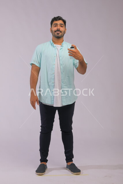 الروائح الطيبة الفواحه ،عطورات خليجية سعودية, مفهوم الاهتمام بالمظهر الخارجي, استخدام الزيوت العطرية الطبيعية, بورتريه لشاب عربي خليجي سعودي يرتدي الزي الكاجوال و يقوم برش العطر وينظر إلى الكاميرا, التسويق لمنتجات العطور, صورة الجسم بالكامل, خلفية رمادية