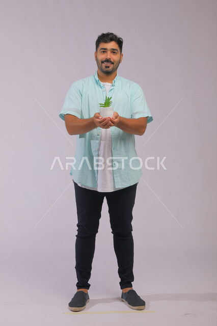 الوقوف باستقامة و النظر إلى الكاميرا بإيماءات السرور ،بورتريه لشاب عربي خليجي سعودي يرتدي الزي الكاجوال ويحمل بيده حوض زراعي صغير ، حب النباتات والاهتمام بها، العناية بالشتلات المنزلية، صورة الجسم بالطول الكامل ،خلفية رمادية