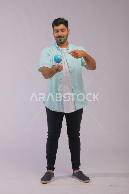 The concept of searching and exploring around the world, pointing to a specific place on the map and looking at one of the continents, portrait of a smiling Saudi Arabian Gulf man wearing casual clothes and holding a globe in his hand, full-length body portrait, gray background