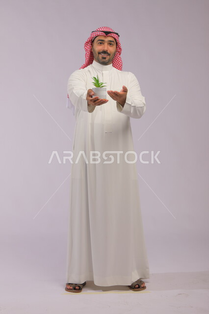 الاهتمام بالشتلات المنزلية, العناية بنباتات الزينة, بورتريه لرجل عربي خليجي سعودي مبتسم يرتدي الثوب التقليدي والشماغ يحمل بيده حوض زراعي صغير, الوقوف باستقامة والنظر إلى الكاميرا بإيماءات السرور والبشاشة, صورة الجسم بالطول الكامل, خلفية رمادية