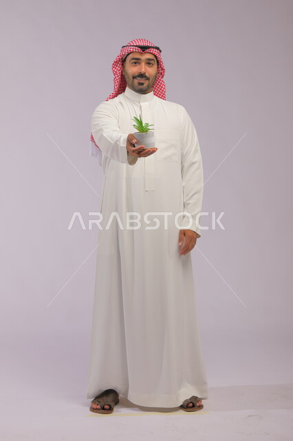 الاهتمام بالشتلات المنزلية, العناية بنباتات الزينة, بورتريه لرجل عربي خليجي سعودي مبتسم يرتدي الثوب التقليدي والشماغ يحمل بيده حوض زراعي صغير, الوقوف باستقامة والنظر إلى الكاميرا بإيماءات السرور والبشاشة, صورة الجسم بالطول الكامل, خلفية رمادية