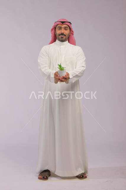 العناية بنباتات الزينة, الاهتمام بالشتلات المنزلية, بورتريه لرجل عربي خليجي سعودي مبتسم يرتدي الثوب التقليدي والشماغ يحمل بيده حوض زراعي صغير, الوقوف باستقامة والنظر إلى الكاميرا بإيماءات السرور والبشاشة, صورة الجسم بالطول الكامل, خلفية رمادية
