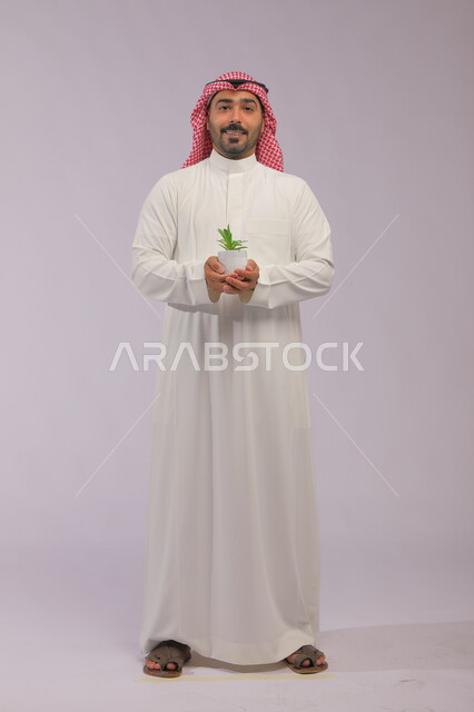 العناية بنباتات الزينة, الاهتمام بالشتلات المنزلية, بورتريه لرجل عربي خليجي سعودي مبتسم يرتدي الثوب التقليدي والشماغ يحمل بيده حوض زراعي صغير, الوقوف باستقامة والنظر إلى الكاميرا بإيماءات السرور والبشاشة, صورة الجسم بالطول الكامل, خلفية رمادية