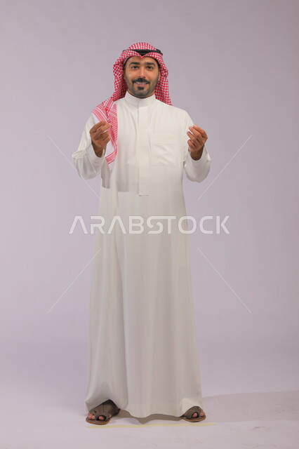  الإشارة بإيماءات طلب الصبر والتأني والتريث، بورتريه لرجل عربي خليجي سعودي يرتدي الزي التقليدي والشماغ, السرور بالصفقات الناجحة والنمو الاقتصادي والمالي, صورة الجسم بالطول الكامل, خلفية رمادية.