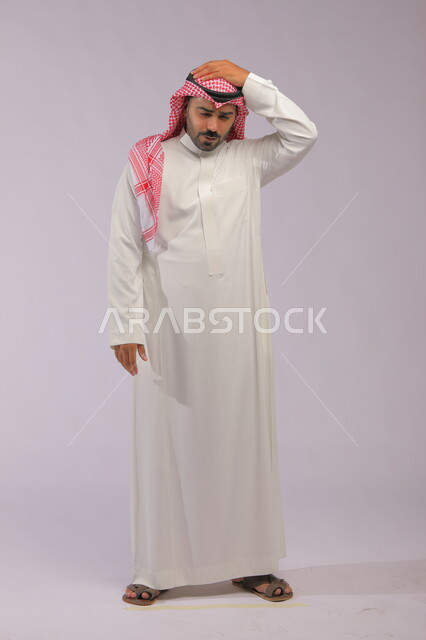  تذكر شيء أو أمر ما، بورتريه لرجل عربي سعودي خليجي يرتدي الثوب السعودي بإيماءات وجه ويدين تدل على الشعور بالصداع وألم بالرأس, التعب والإرهاق والإحساس بالمرض, آلام الشقيقة والصداع النصفي, خلفية رمادية.