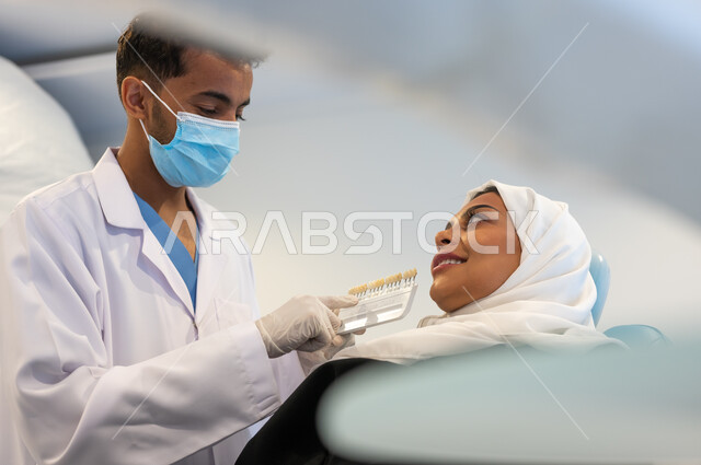 مفهوم الرعاية والاهتمام بصحة ومظهر الأسنان، طبيب أسنان عربي خليجي سعودي يرتدي القفازات والكمامة الطبية يمسك بيده دليل الألوان لترميم أسنان المريضة، سيدة عربية خليجية سعودية في عيادة سنية تتلقى العلاج اللازم لصنع ابتسامة مميزة.