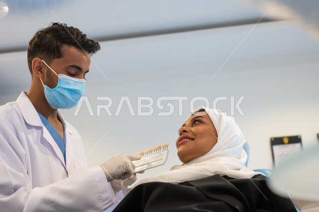 مفهوم الرعاية والاهتمام بصحة ومظهر الأسنان، طبيب أسنان عربي خليجي سعودي يرتدي القفازات والكمامة الطبية يمسك بيده دليل الألوان لترميم أسنان المريضة، سيدة عربية خليجية سعودية في عيادة سنية تتلقى العلاج اللازم لصنع ابتسامة مميزة.