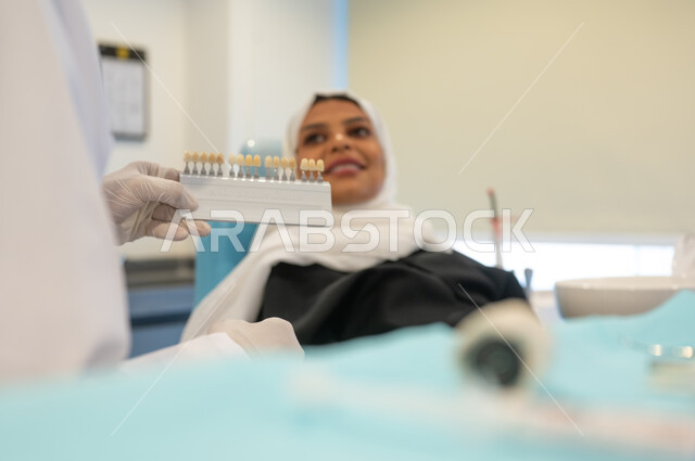 تقديم الخدمات والرعاية الصحية في المراكز والعيادات التخصصية، طبيب أسنان عربي خليجي سعودي يرتدي المعطف والقفازات والكمامة الطبية يمسك بيده دليل الألوان لترميم أسنان المريضة، سيدة عربية خليجية سعودية في عيادة سنية تتلقى العلاج اللازم للحصول على ابتسامة مثالية.