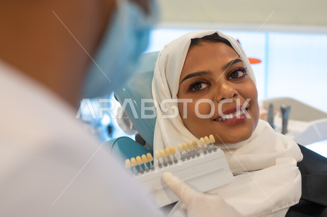 Taking care of the appearance of teeth to get a perfect smile, a close-up picture of a Saudi Gulf Arab dentist wearing a coat, gloves and a medical mask holding in his hand the color guide for restoring the patient’s teeth, a veiled Saudi Gulf Arab woman in a dental clinic receiving the necessary treatment.