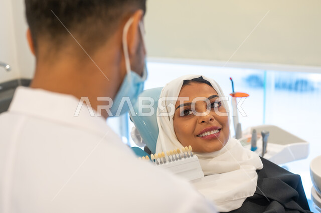 Taking care of the appearance of teeth to get a perfect smile, a close-up picture of a Saudi Gulf Arab dentist wearing a coat, gloves and a medical mask holding in his hand the color guide for restoring the patient’s teeth, a veiled Saudi Gulf Arab woman in a dental clinic receiving the necessary treatment.