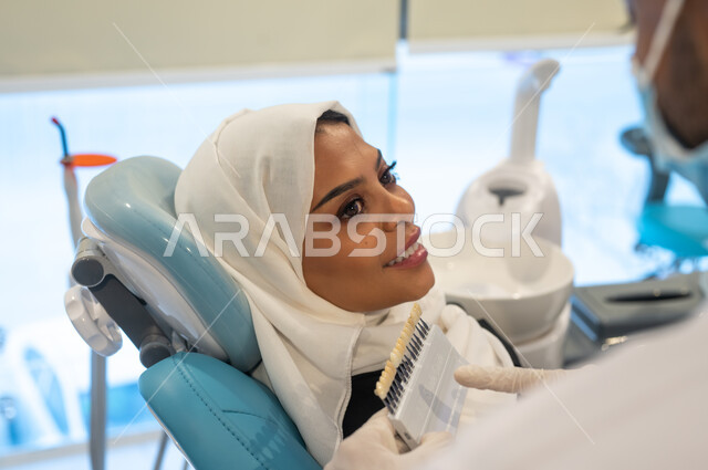 العناية بمظهر الأسنان للحصول على ابتسامة مثالية، صورة مقربة لطبيب أسنان عربي خليجي سعودي يرتدي المعطف والقفازات والكمامة الطبية يمسك بيده دليل الألوان لترميم أسنان المريضة، سيدة عربية خليجية سعودية محجبة في عيادة سنية تتلقى العلاج اللازم.