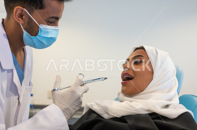 العناية بالفم بالطرائق العلاجية الصحيحة، حقن البنج الموضعي لتخفيف الإحساس بالألم، صورة مقربة طبيب أسنان عربي خليجي سعودي يرتدي المعطف والقفازات والكمامة الطبية يمسك بيده إبرة التخدير، سيدة عربية خليجية سعودية محجبة في عيادة سنية تتلقى العلاج اللازم.