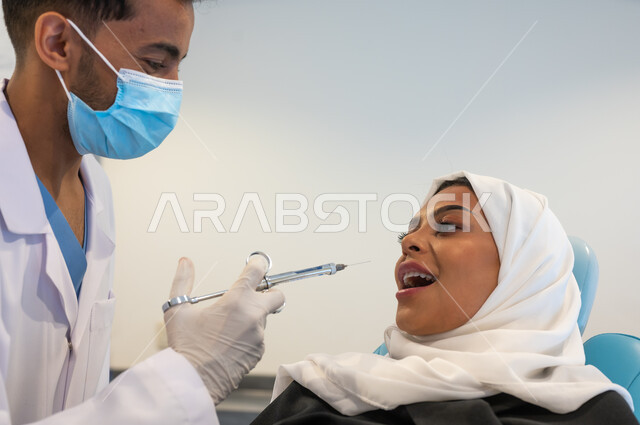 العناية بالفم بالطرائق العلاجية الصحيحة، حقن البنج الموضعي لتخفيف الإحساس بالألم، صورة مقربة طبيب أسنان عربي خليجي سعودي يرتدي المعطف والقفازات والكمامة الطبية يمسك بيده إبرة التخدير، سيدة عربية خليجية سعودية محجبة في عيادة سنية تتلقى العلاج اللازم.