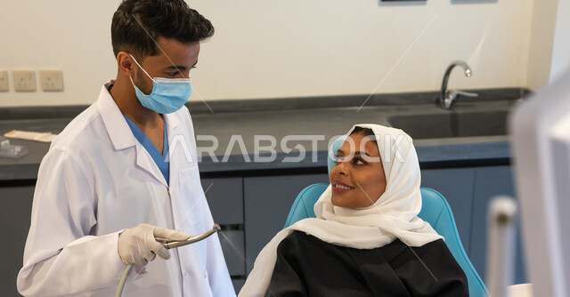 Visit health clinics and take the necessary measures. A Saudi Gulf Arab dentist wearing a coat, gloves, and a medical mask holds in his hand a tooth drilling tool and removing cavities. A veiled Saudi Gulf Arab woman in a dental clinic receiving the necessary treatment.