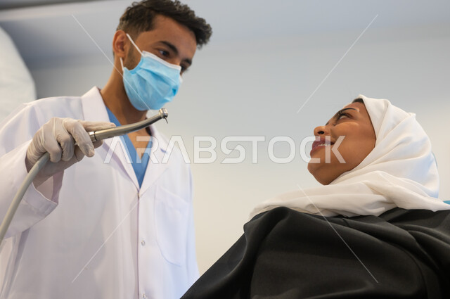 معدات وأدوات طب الأسنان، زيارة العيادات الصحية واتخاذ الإجراءات اللازمة، طبيب أسنان عربي خليجي سعودي يرتدي المعطف والقفازات والكمامة الطبية يمسك بيده أداة حفر السن وإزالة التسوس، سيدة عربية خليجية سعودية محجبة في عيادة سنية.