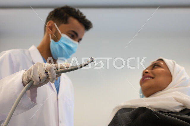 معدات وأدوات طب الأسنان، زيارة العيادات الصحية واتخاذ الإجراءات اللازمة، طبيب أسنان عربي خليجي سعودي يرتدي المعطف والقفازات والكمامة الطبية يمسك بيده أداة حفر السن وإزالة التسوس، سيدة عربية خليجية سعودية محجبة في عيادة سنية.