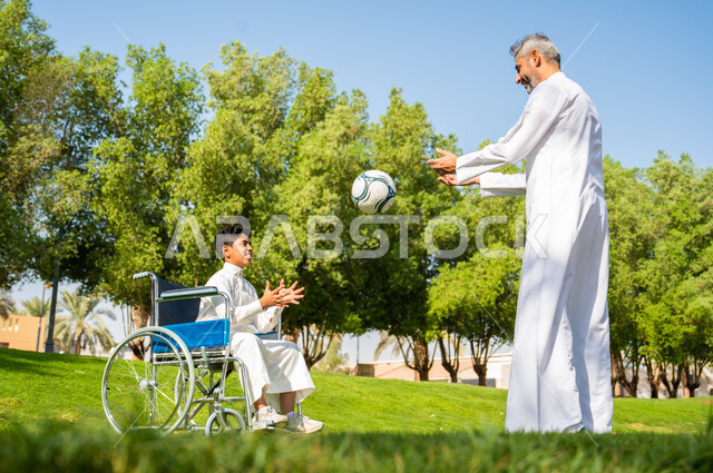 Paying attention to the psychological and physical aspects of people with disabilities, gestures of cheerfulness and good treatment, a Saudi Arabian Gulf father wearing traditional clothing playing with his son of special needs, sitting in a wheelchair and spending time in the park with the family, amusement and entertainment playing with the ball, nature background