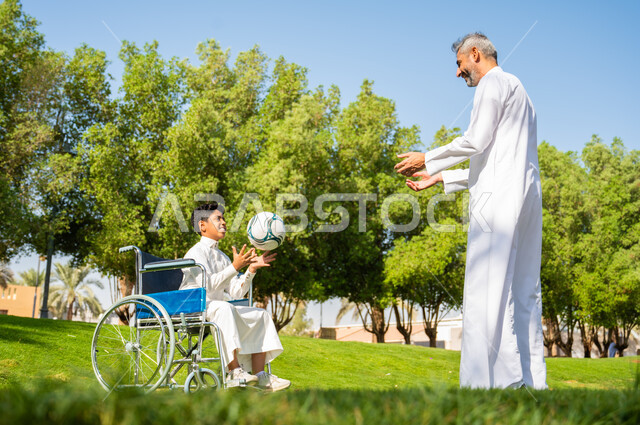 التسلية والترفيه في اللعب مع الأبناء بالكرة، إيماءات البشاشه والمعاملة الحسنة, أب عربي سعودي خليجي يرتدي الزي التقليدي يلعب مع ولده من ذوي الهمم، الجلوس على كرسي متحرك وقضاء الوقت في المنتزه, الاهتمام بالجانب النفسي والجسدي للمقعدين, خلفية الطبيعة.
