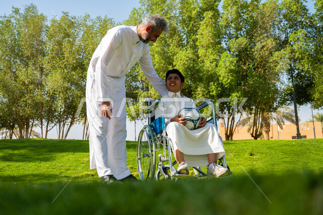 إيماءات البشاشه والمعاملة الحسنة, أب عربي سعودي خليجي يرتدي الزي التقليدي يتحادث مع ابنه من ذوي الهمم، الجلوس على كرسي متحرك وقضاء الوقت في المنتزه مع العائلة, اهتمام ودعم المجتمع لذوي الاحتياجات الخاصه, الاستعداد للعب في الكرة، خلفية الطبيعة. 