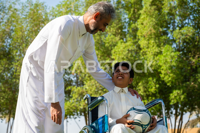 قضاء الوقت في المنتزه مع الأبناء, أب يتبادل الأحاديث مع ابنه المقعد, ولد عربي سعودي خليجي من ذوي الهمم  يرتدي الزي التقليدي يجلس على كرسي متحرك و يمسك بيده الكرة, اهتمام ودعم المجتمع لذوي الاحتياجات الخاصه, خلفية الطبيعة.