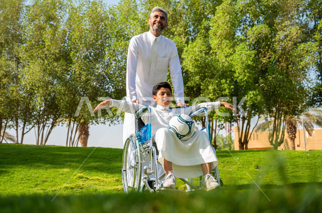 A father offering love, care and attention to his disabled son, a Saudi Arabian Gulf boy with special needs wearing traditional clothing, sitting in a wheelchair and holding a ball in his hand, spending time in the park with the family, community interest and support for people with special needs, nature background