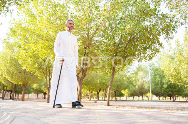 الخروج إلى أحد الحدائق العامة للترفيه عن النفس، نزهه في الطبيعه و الهواء الطلق، الاستمتاع بالأجواء الصيفيه في إحدى المنتزهات، رجل كبير السن عربي خليجي سعودي يرتدي الثوب التقليدي يرتكز على عكاز لسهولة التنقل بإيماءات السعادة، معدات تساعد على المشي