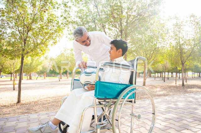 قضاء الوقت في المنتزه مع العائلة ،العطف والحنان بين أفراد الأسرة ،مفهوم العنايه ورعاية المرضى والمعاقين، أب يقوم بالاهتمام بالجانب النفسي والجسدي لابنه, ولد عربي سعودي خليجي من ذوي الهمم يرتدي الزي التقليدي يجلس على كرسي متحرك و يمسك بيده الكرة