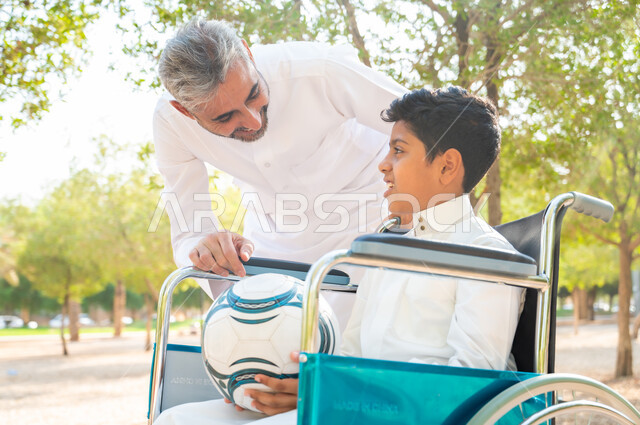 مفهوم العنايه ورعاية المرضى والمعاقين, أب يقوم بالاهتمام بالجانب النفسي والجسدي لابنه, ولد عربي سعودي خليجي من ذوي الهمم يرتدي الزي التقليدي يجلس على كرسي متحرك و يمسك بيده الكرة, قضاء الوقت في المنتزه مع العائلة, العطف والحنان بين أفراد الأسرة