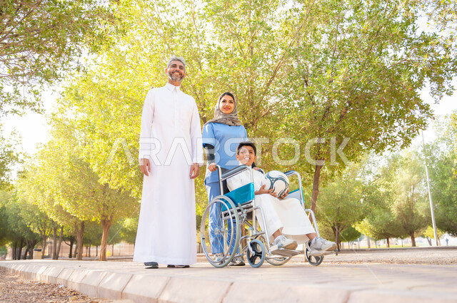 مفهوم الرعاية والعناية بالمرضى والمعاقين، قضاء الإجازة في المنتزه مع العائلة ،العطف والحنان بين أفراد الأسرة، ممرضة سعودية تقوم بمساعدة مريضها والاهتمام به, ولد عربي سعودي خليجي من ذوي الهمم يرتدي الزي التقليدي يجلس على كرسي متحرك برفقة والده في الخارج
