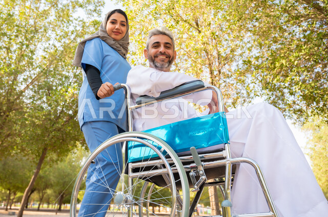 قوة الارادة و العزيمة عند أصحاب الهمم, النظر إلى الكاميرا بإيماءات السرور، شعور المعاقين بالإنتماء والتقدير والإحترام في السعوديه ،رجل كبير السن عربي خليجي سعودي مبتسم مقعد يرتدي الثوب التقليدي يجلس على كرسي متحرك برفقة ممرضته الخاصة في إحدى المنتزهات