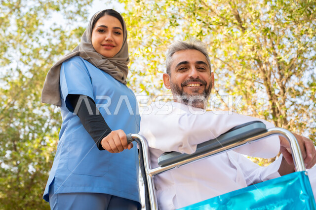 النظر إلى الكاميرا بإيماءات السرور، شعور المعاقين بالإنتماء والتقدير والإحترام في السعوديه ،قوة العزيمة والتحدي عند أصحاب الهمم, صورة مقربة لرجل مسن عربي خليجي سعودي مبتسم مقعد يرتدي الثوب التقليدي يجلس على كرسي متحرك برفقة ممرضته الخاصة في إحدى الحدائق العامة