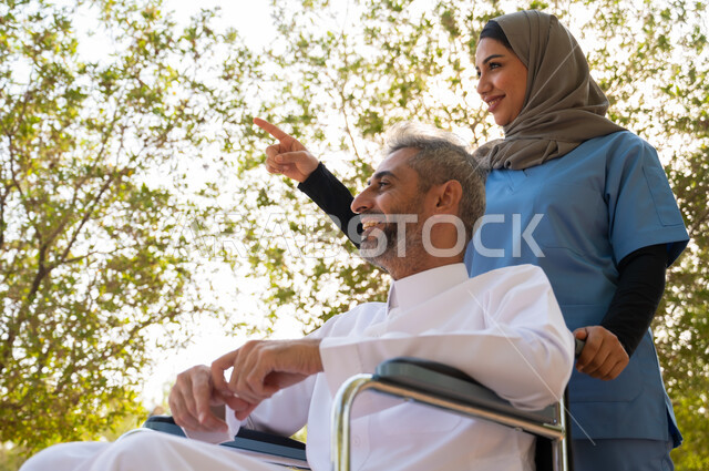 الاشارة إلى شيء ما بإيماءات السرور، شعور المعاقين بالإنتماء والتقدير والإحترام في السعوديه ،قوة العزيمة والتحدي عند أصحاب الهمم, صورة مقربة لرجل مسن عربي خليجي سعودي مبتسم مقعد يرتدي الثوب التقليدي يجلس على كرسي متحرك برفقة ممرضته الخاصة في إحدى الحدائق العامة