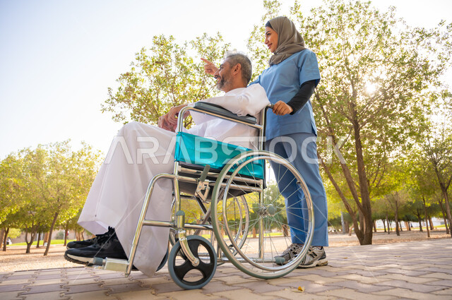 مفهوم العنايه ورعاية المرضى والمعاقين ،قوة العزيمة والتحدي عند أصحاب الهمم, صورة مقربة لرجل كبير السن عربي خليجي سعودي مبتسم مقعد يرتدي الثوب التقليدي يجلس على كرسي متحرك برفقة ممرضته الخاصة في إحدى الحدائق العامة والاشارة إلى شيء ما