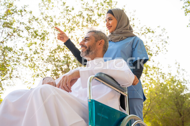 The feeling of belonging, appreciation and respect for people with disabilities in Saudi Arabia, the strength of determination and challenge among people of determination, a close-up picture of an elderly smiling Saudi Gulf Arab man wearing a traditional dress sitting in a wheelchair with his private nurse in a public park and pointing to something